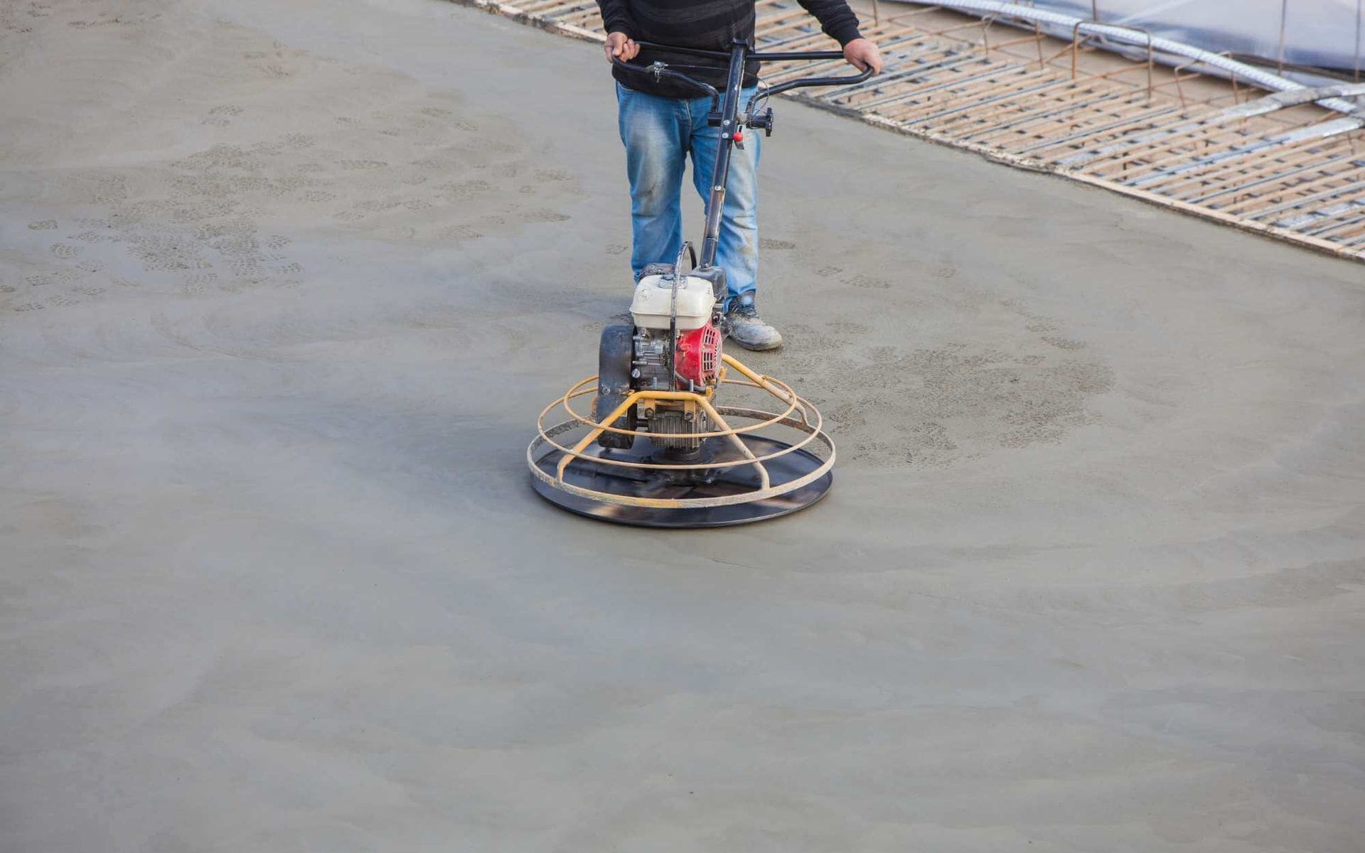 A construction worker in Buckeye AZ smoothens a concrete surface using a power trowel. Wearing jeans and holding the machine's handles firmly, he guides it over the freshly laid decorative concrete. Construction material is visible in the background, showcasing the expertise of local concrete contractors.