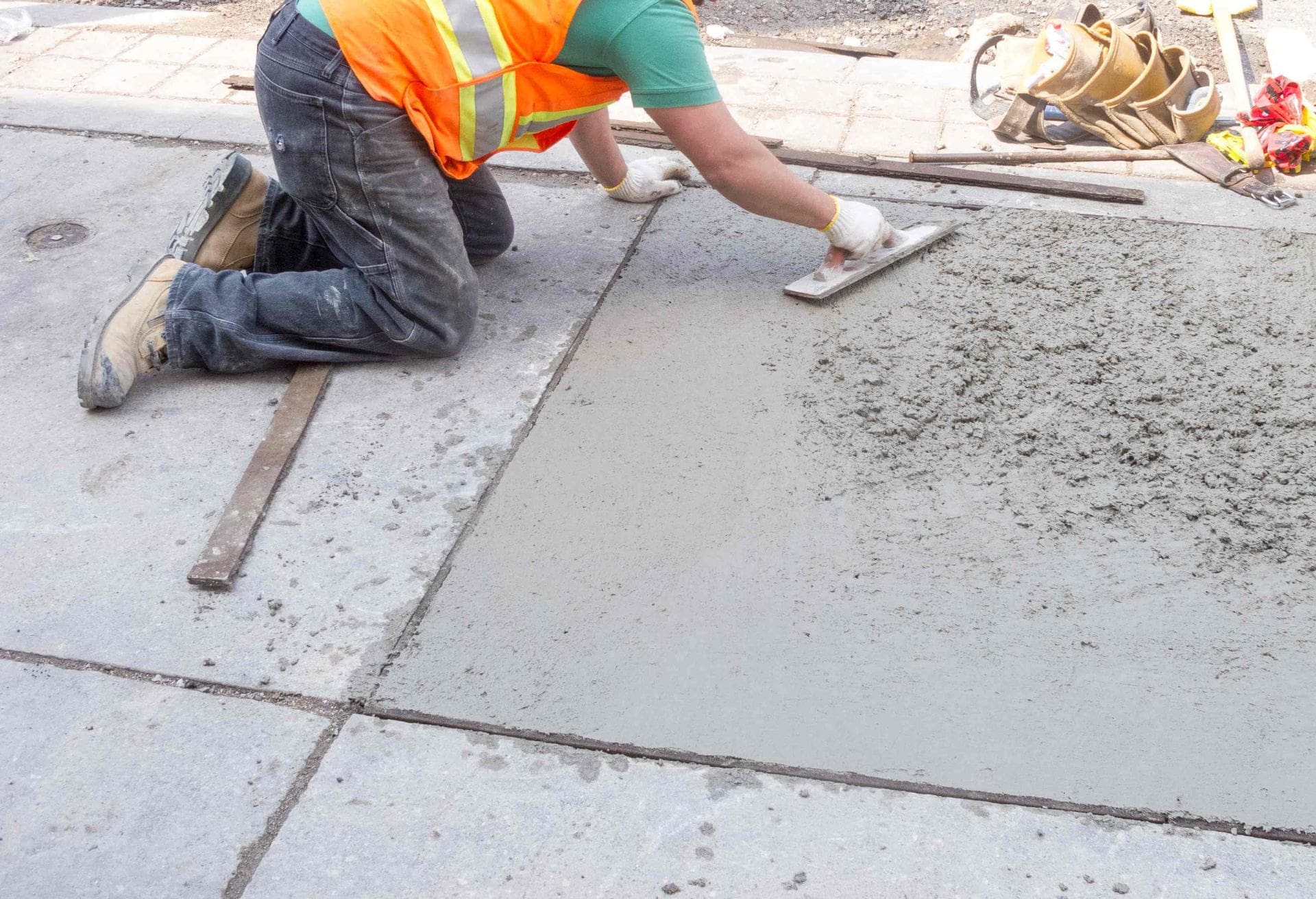 A construction worker wearing an orange safety vest and kneeling on the ground smooths wet concrete on a Sun City sidewalk with a trowel. Wooden planks outline the area being worked on, and West Valley Concrete materials are visible in the background.
