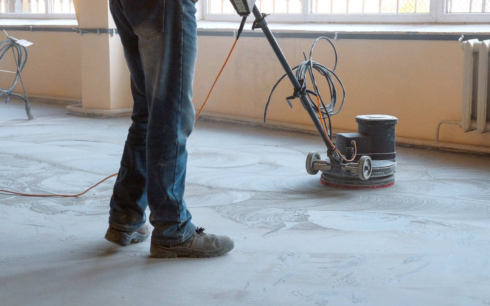 A person is using a floor buffer to polish a concrete floor. They are wearing jeans and work boots. Electric cables connect the floor buffer, and dust is visible on the floor around the machine. The space has beige walls and minimal furniture, reflecting the quality work of a skilled concrete company in Surprise AZ.