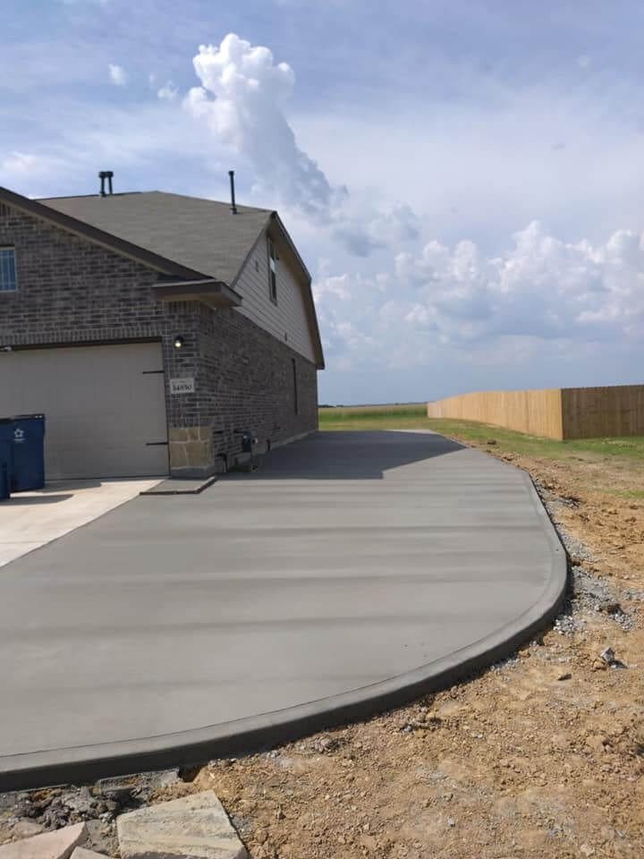 A newly paved concrete driveway, a testament to high-quality concrete results, curves around the side of a brick house on a sunny day. The driveway leads to a two-car garage, with a wooden fence standing along the right edge of the property. The sky is filled with fluffy clouds.