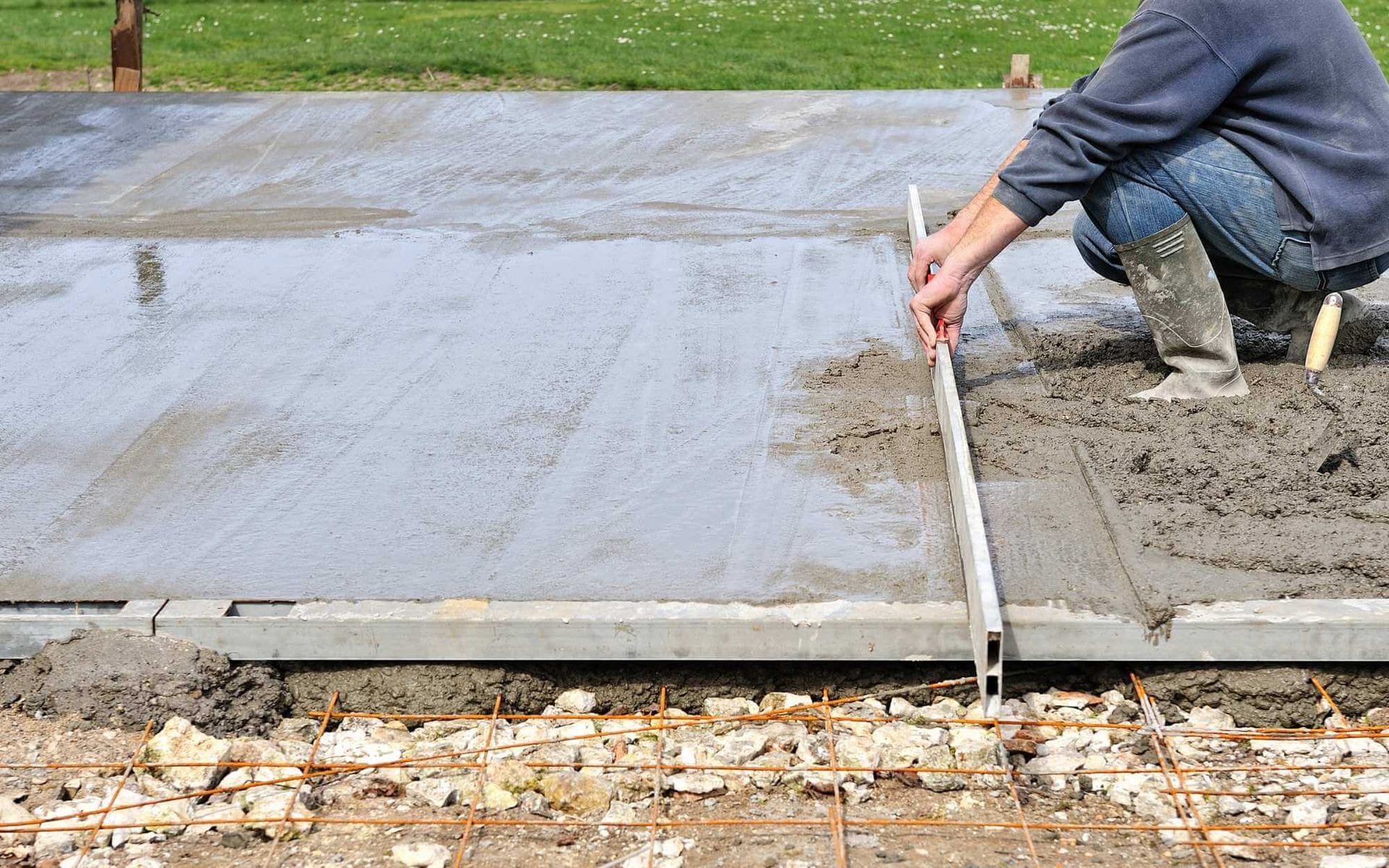 A person is using a long, straight board to level freshly poured concrete on a construction site in El Mirage. Wearing a gray sweatshirt and rubber boots, they ensure the concrete meets West Valley Concrete standards. The mix smooths out, with some wire mesh and rocks visible at the edge of the pour.
