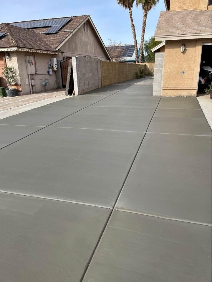 A clean, newly paved concrete driveway stretches from the garage of a house to the street, crafted with precision by West Valley Concrete services. Bordered by a beige wall on one side and an adjacent home on the other, it sits in the sunny charm of Sun City. Solar panels shine on both roofs as palm trees sway in the background.