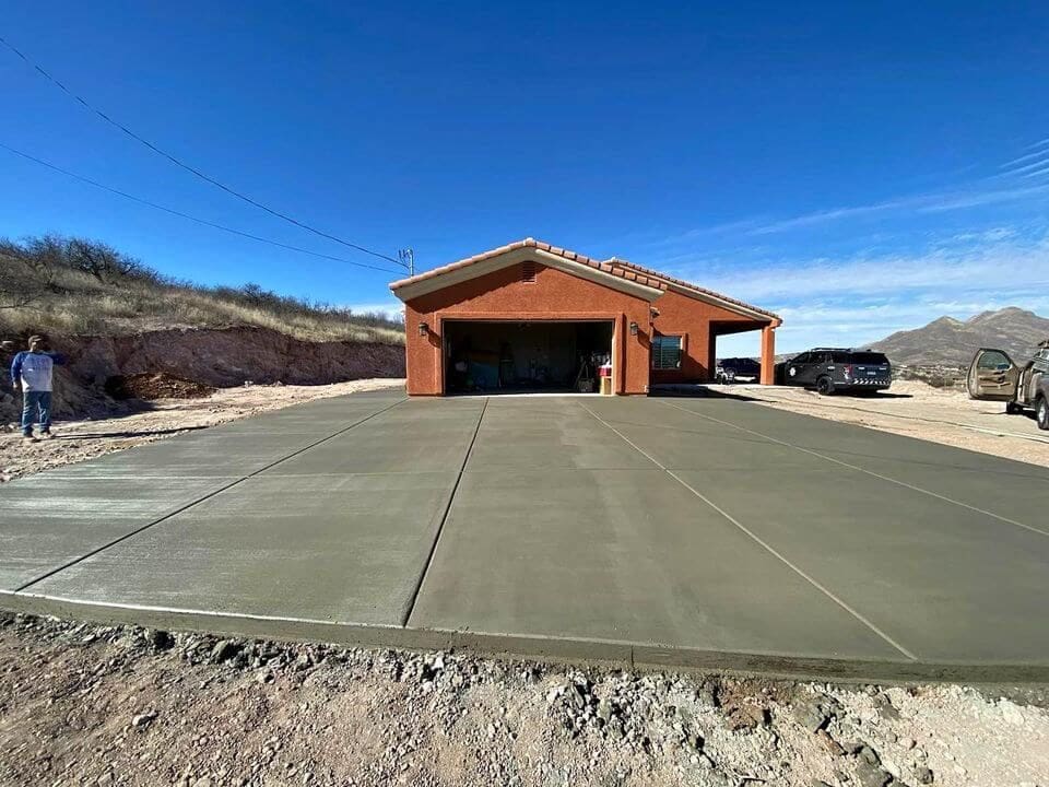 A newly poured concrete driveway by West Valley Concrete leads to a single-story brick house with an open garage. Two people stand on the left side, and two vehicles are parked on the right. The rugged terrain with hills is bathed in sunlight under a clear blue sky.