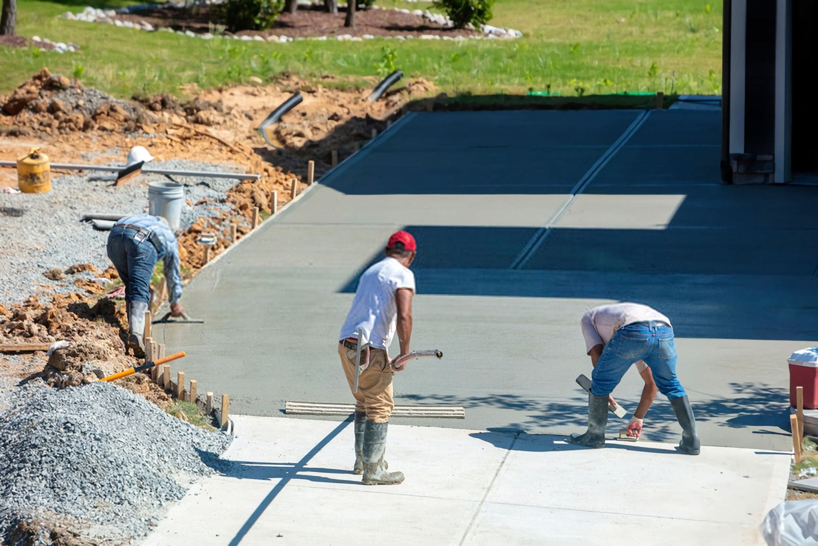 Three construction workers wearing casual clothing and safety boots are diligently smoothing and finishing the surface of a nearly completed concrete driveway. Construction material and debris are visible around the area, showcasing the efficient work of a trusted concrete contractor in Glendale AZ.