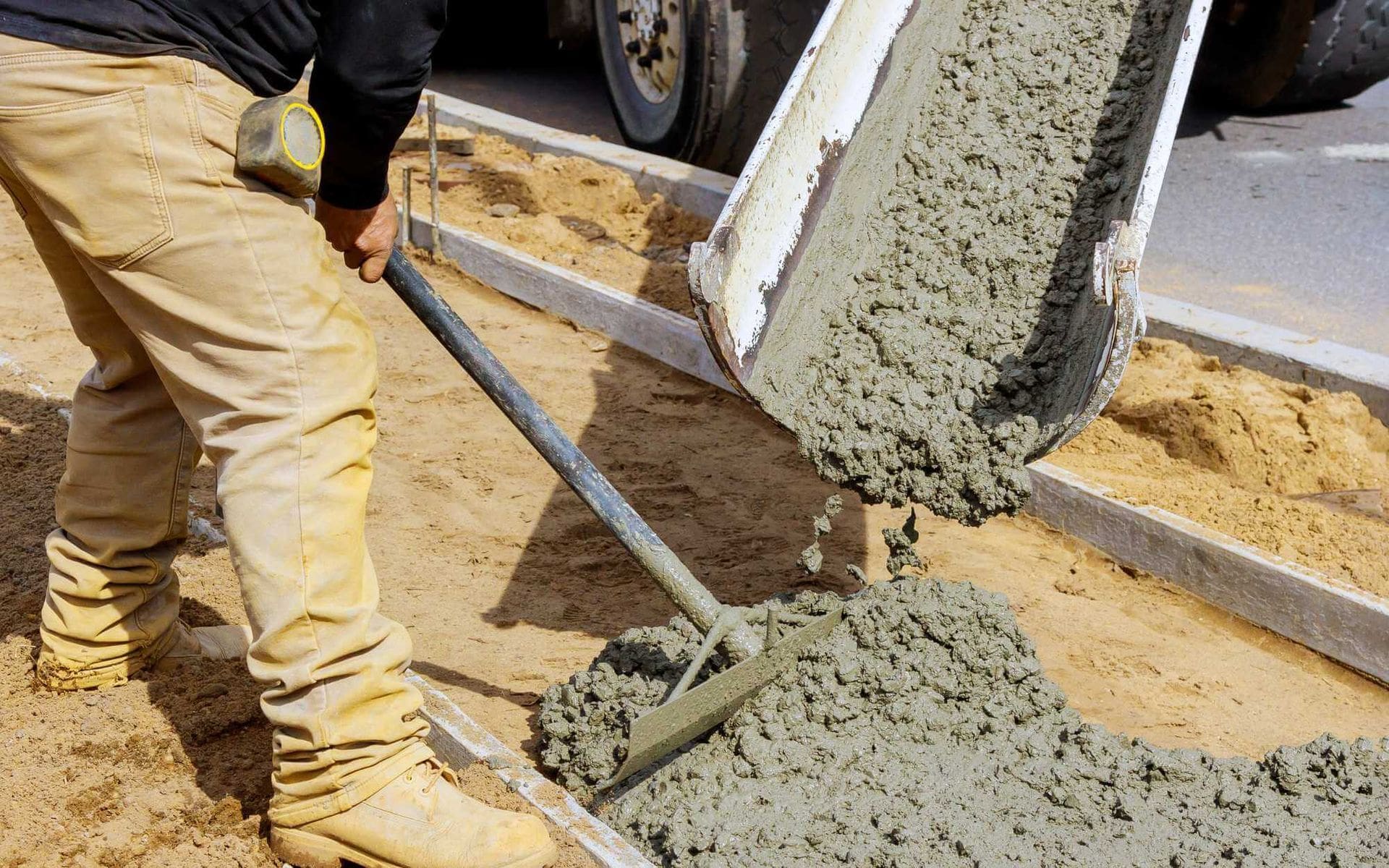 A construction worker wearing beige pants and boots is spreading fresh concrete with a rake. The concrete is being poured from a chute into a rectangular formwork on the ground. Sand surrounds the work area as skilled concrete contractors in Buckeye, AZ ensure flawless application, possibly for decorative concrete.