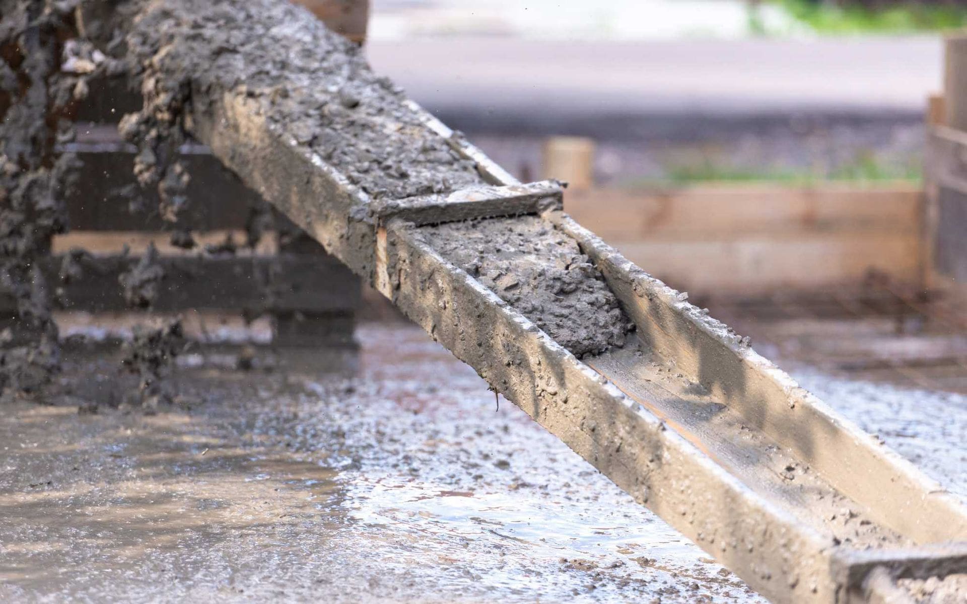 A close-up view of wet concrete being poured down a metal chute at a West Valley Concrete construction site. The surrounding area is blurred with a mix of water and concrete on the ground, indicating active construction work. Contact us today for a free quote to meet all your concrete needs.