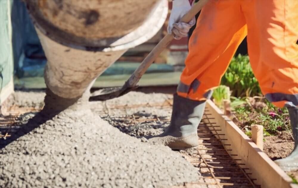 A construction worker in orange pants and black boots spreads freshly poured concrete from a West Valley Concrete mixer truck onto a metal rebar grid at a building site.