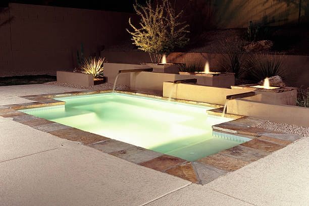 A modern, illuminated rectangular pool with cascading water features is set against a nighttime backdrop. The pool is surrounded by a light-colored stone deck, built by a top pool deck contractor in Glendale, AZ. Built-in steps and meticulously landscaped small plants are enhanced by strategic lighting.