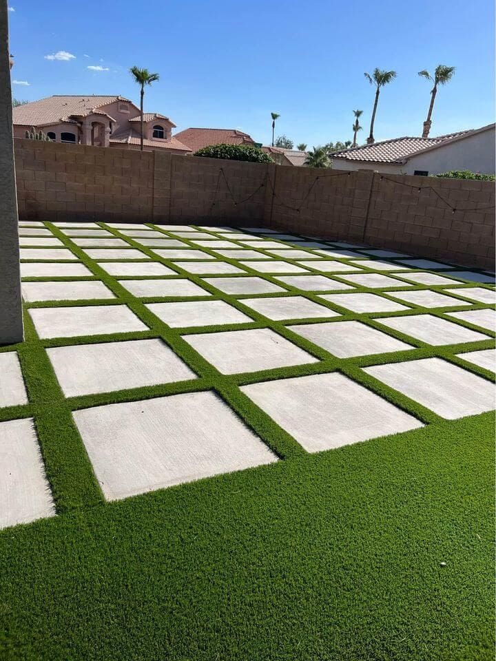 A backyard with a modern design featuring large concrete squares arranged in a grid pattern, surrounded by green artificial grass. The yard, crafted by West Valley Concrete, is enclosed by a beige brick wall with neighboring houses in the background under a clear blue sky. Get your free quote today!