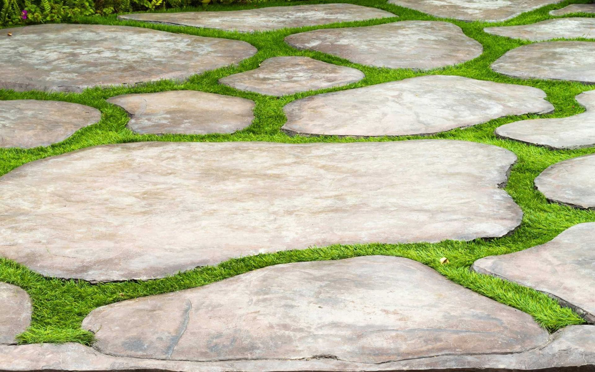A garden walkway in Sun City features large, irregularly shaped stone slabs interspersed with vibrant green grass, creating a natural and organic pattern. The stones vary in size and shape, with grass filling the gaps between them. For similar designs, consider West Valley Concrete services for expert craftsmanship.