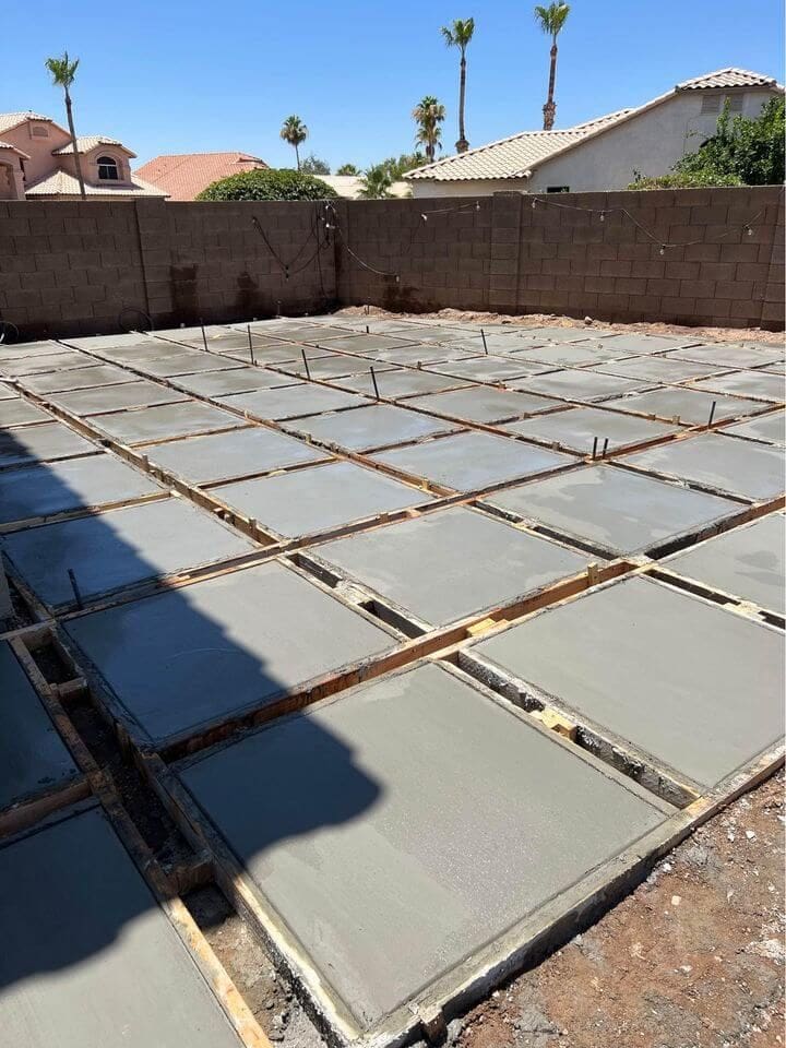 Freshly poured concrete slabs within wooden frames are set on the ground, forming a grid pattern. A brick wall and houses with tiled roofs are in the background. Some metal rods are protruding from the concrete, under a clear blue sky—work expertly done by your reliable concrete company offering free quotes in Surprise AZ.