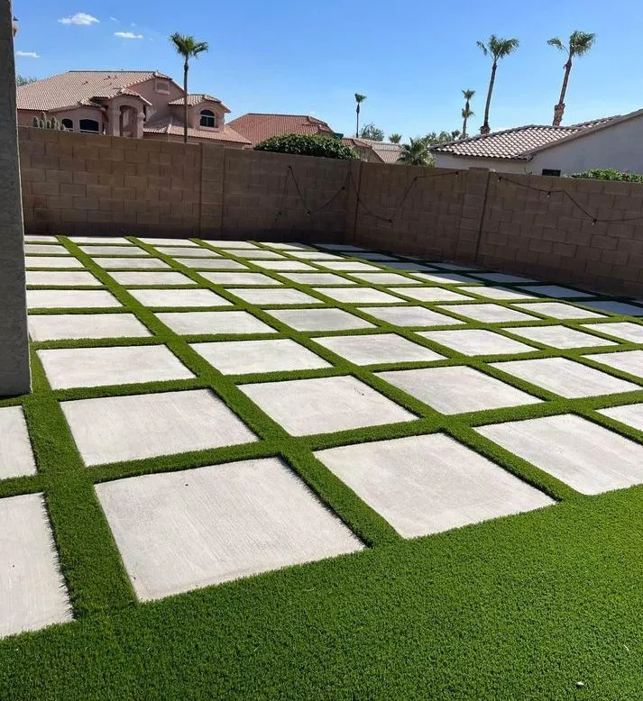 A backyard in Peoria, Arizona, with a modern design featuring a decorative concrete grid pattern of square pavers interspersed with artificial grass. The yard is enclosed by a beige brick wall, and palm trees and neighboring houses are visible in the background under a clear blue sky.