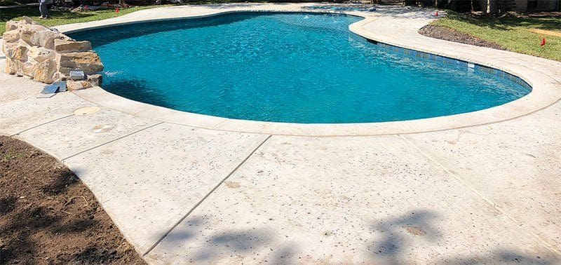 A backyard swimming pool with clear blue water is surrounded by a light-colored concrete deck, expertly crafted by a licensed and insured Glendale AZ pool deck contractor. A small rock formation graces the corner of the pool, with some grass and landscaping visible in the background. Get a free quote today!