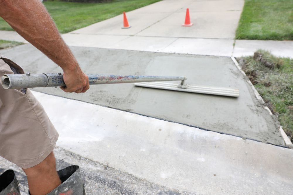 A person smooths wet concrete on a sidewalk using a long-handled trowel, a sign of meticulous concrete resurfacing. Two orange traffic cones are placed in the background on the adjacent road, indicating an area under construction. Grass lines the edges of the sidewalk.