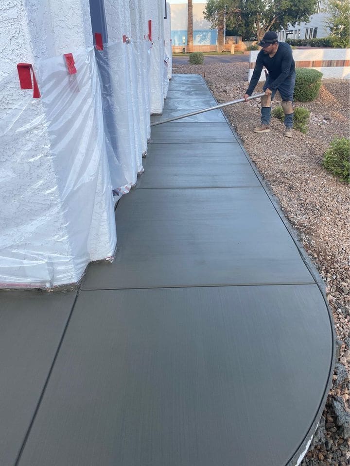 A worker is smoothing freshly poured concrete on a sidewalk alongside a building partially wrapped in protective plastic sheeting. Surrounding the sidewalk, there is gravel landscaping and some shrubs in the background, hinting at a potential patio upgrade or even concrete resurfacing for improved aesthetics.