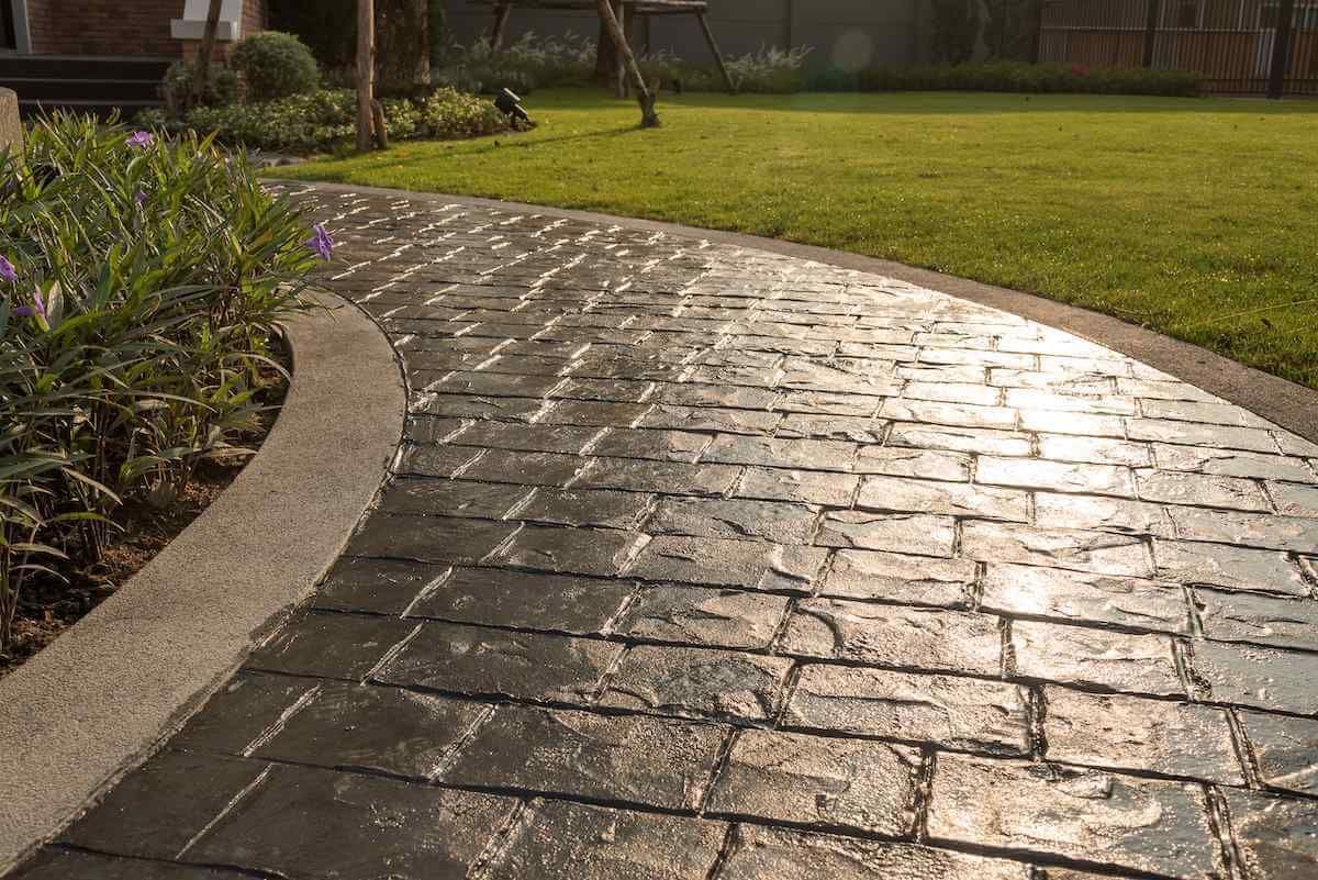 A sunlit, wet cobblestone pathway curves around a landscaped garden with neatly trimmed grass and a flower bed. The stones glisten in the light, showcasing high-quality concrete results. A tree stands in the background, adding to the tranquil outdoor setting.