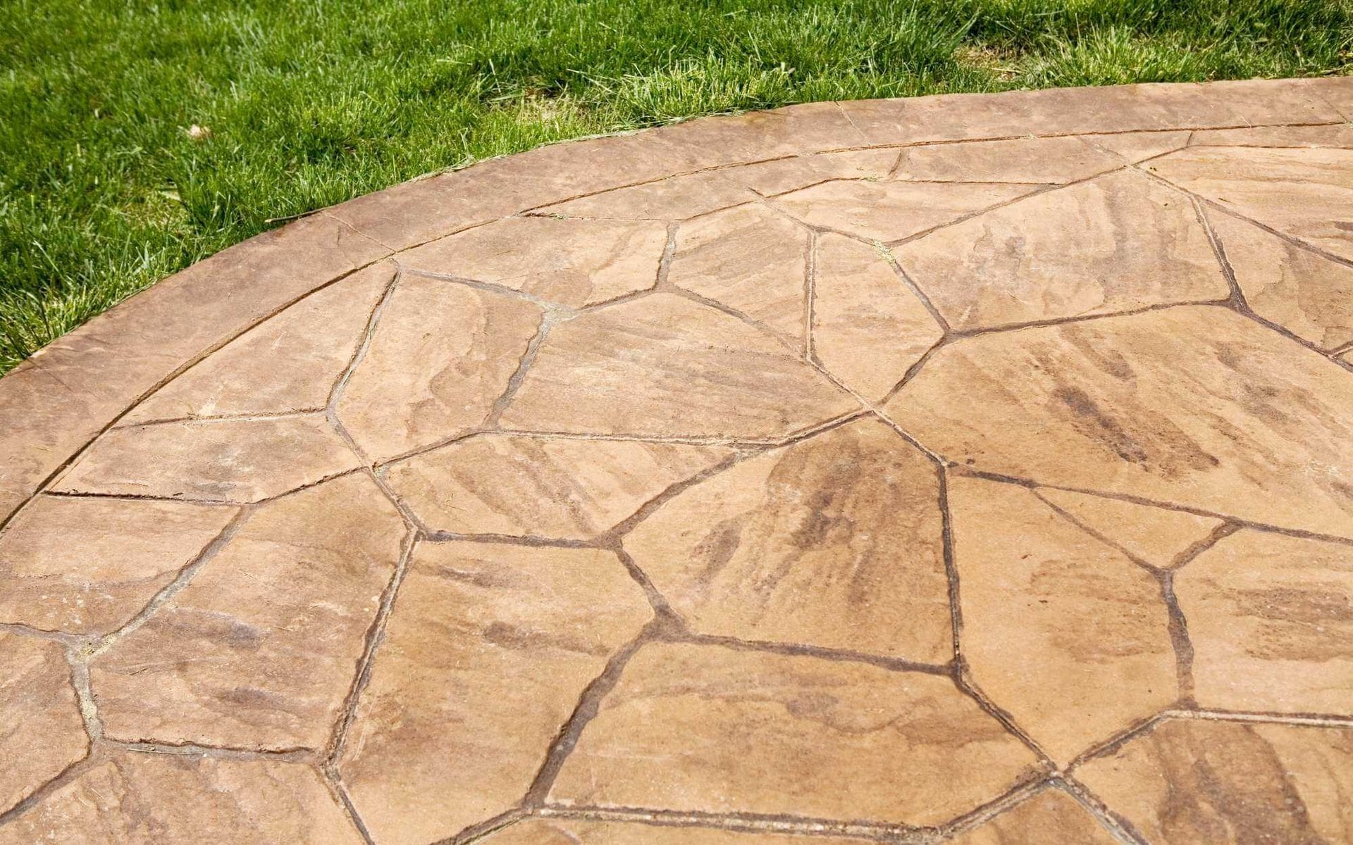 A section of a patio in Peoria, Arizona, made of irregularly shaped, tan-colored flagstone arranged in a circular pattern. The stone surface shows varied textures, with surrounding green grass visible at the top edge of the image. Contact us today for free quotes on all decorative concrete projects!