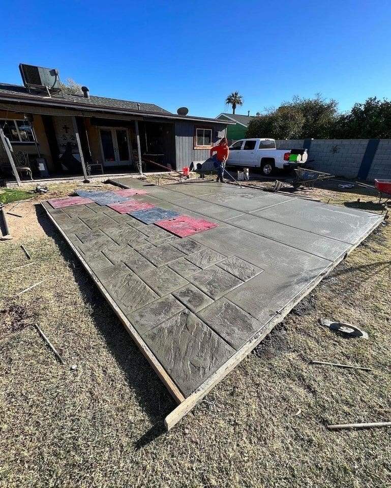A person is working on a partially completed concrete patio in a backyard in Sun City, AZ. The patio features a stamped brick pattern with some sections still wet and colored with red and blue mats. The scene includes a house with a deck, a white pickup truck, and a clear blue sky. Contact us for concrete services and free quotes.