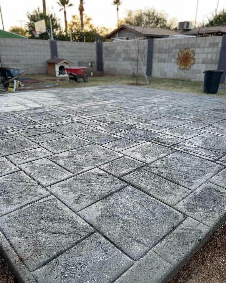 A newly installed stamped concrete patio with a textured stone pattern is shown in a backyard. In the background, there is a cinder block wall, a wheelbarrow, a basketball hoop, and some garden items. Palm trees can be seen beyond the wall. For such stunning results in Surprise AZ concrete projects, trust our reliable concrete company offering free quotes.