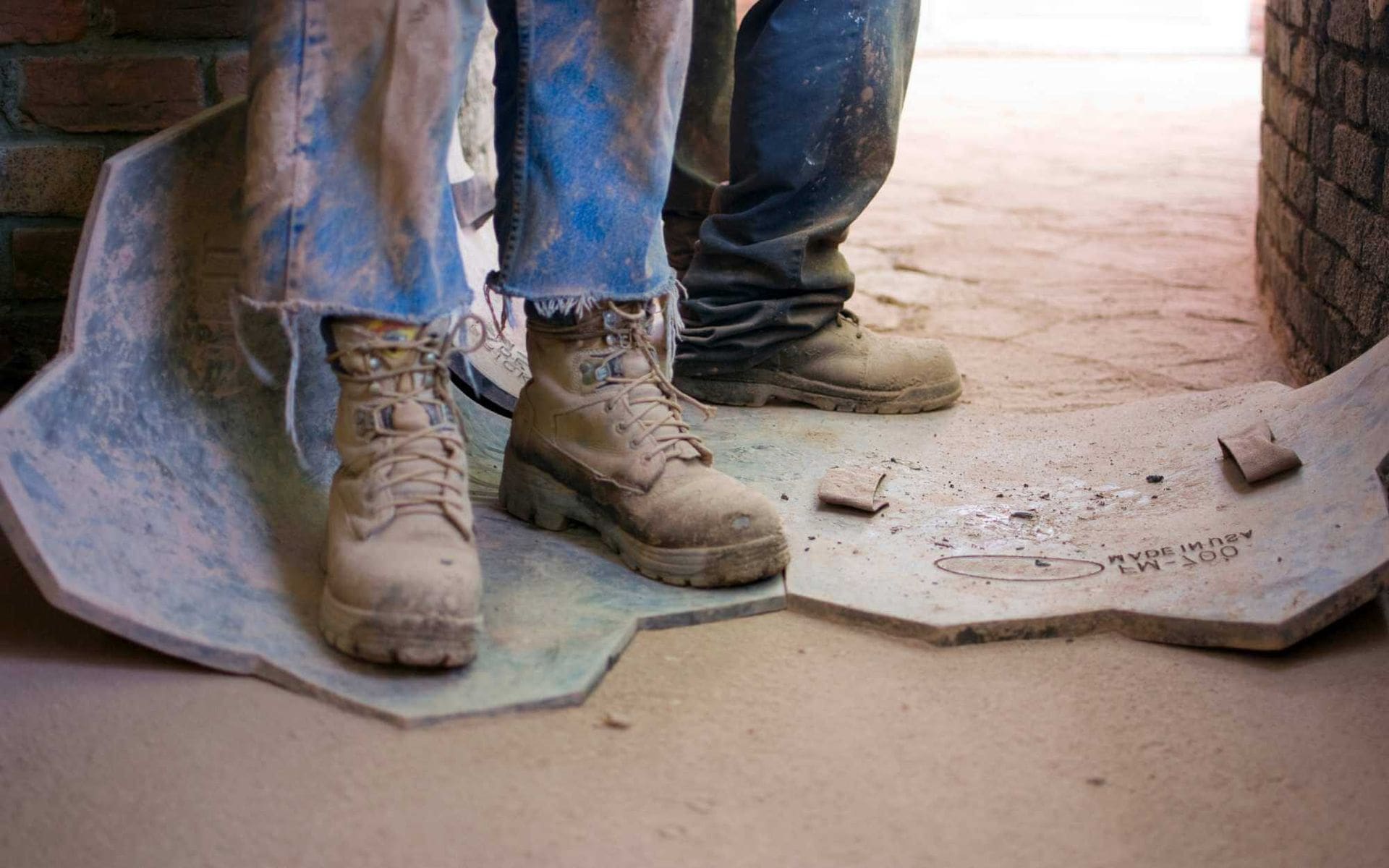 Two people standing on worn-out construction site floor mats in Avondale, AZ. Both are wearing rugged work boots and dirty jeans, with only their feet and lower legs visible. The ground is dusty and cluttered with small debris, reflecting a work-in-progress environment for concrete contractors.