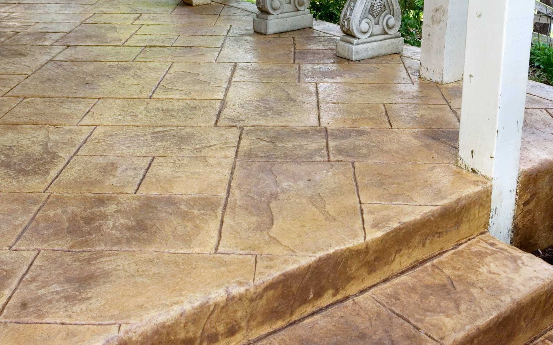 A close-up of a porch in El Mirage with textured, stamped concrete in a pattern resembling large tiles. The concrete has a warm, earthy tone. Decorative stone columns and a bit of foliage are visible in the background, enhancing the aesthetic appeal similar to stylish residential driveways.