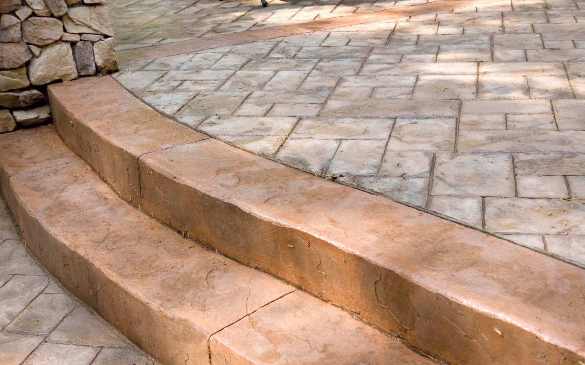 The image shows a close-up of two curved concrete steps with a stamped, textured surface, leading up to a stone wall. The steps and the ground have a similar pattern that resembles large cobblestones or pavers. This decorative concrete installation, sunlit in Peoria Arizona, highlights an outdoor setting.
