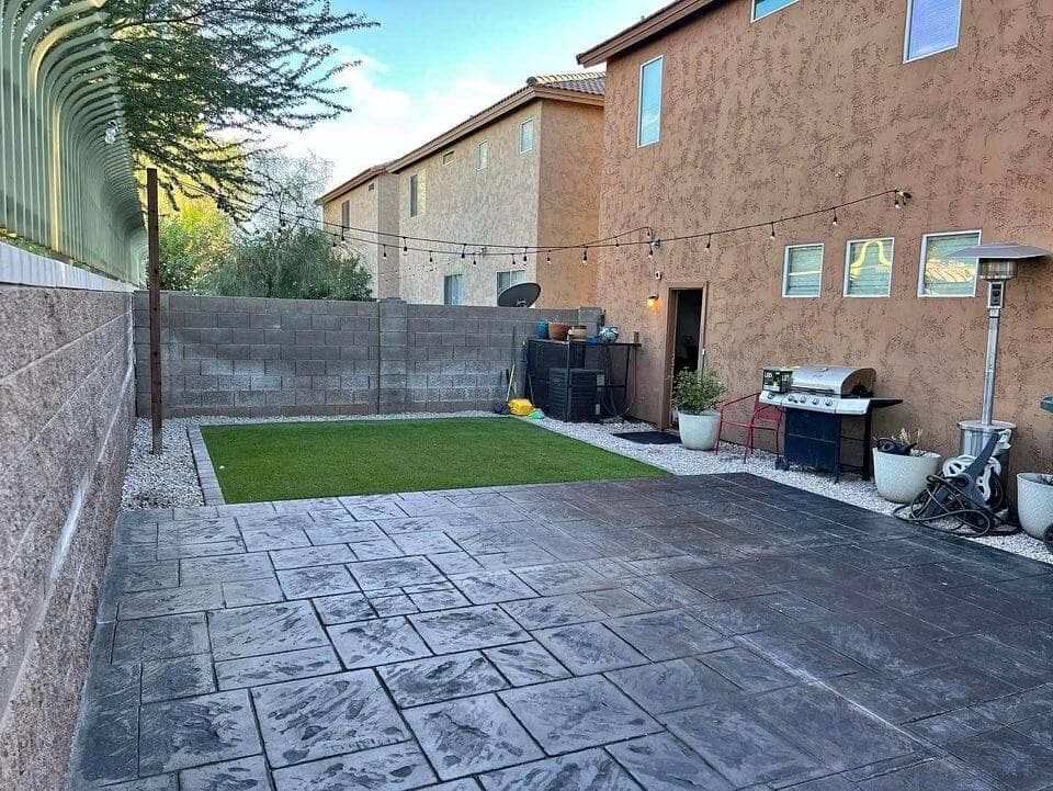 A cozy backyard in Sun City AZ features a tiled patio, a small patch of artificial grass, string lights overhead, and various plants in pots along the perimeter. A barbecue grill and outdoor heater are near a door on the right, set against the brown stucco wall of the house. Contact us for free quotes on concrete services.