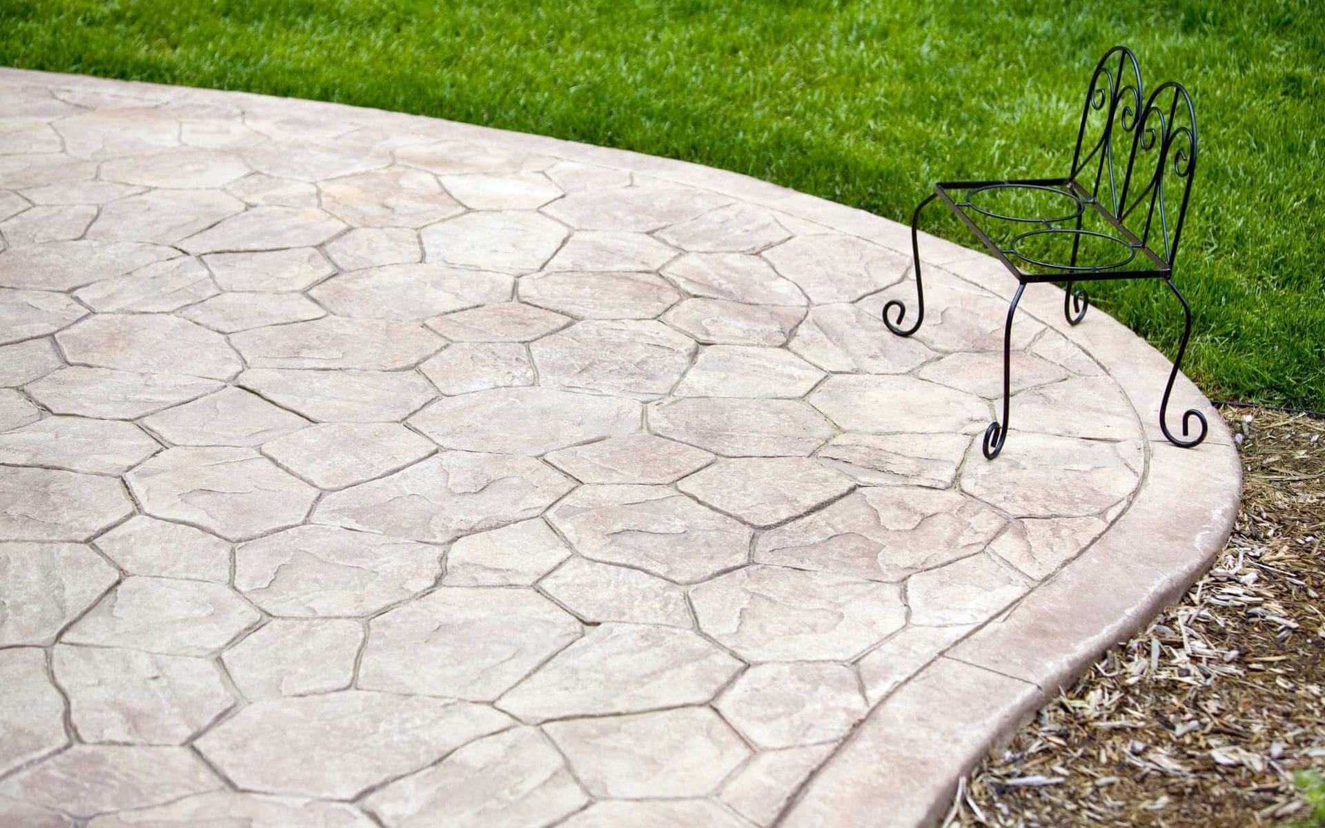 A lone wrought iron chair sits on a light-colored, hexagonal stone patio crafted by West Valley Concrete. The edge of the patio curves gently, meeting a lush green lawn. The simple, elegant design of the chair contrasts with the organic pattern of the patio stones from this reliable concrete company.