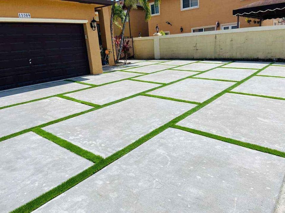 A modern driveway designed by a top concrete contractor in Glendale AZ features large, square concrete slabs with neatly trimmed grass in between each slab. The driveway is adjacent to a garage with a dark door and a light brown house. Small palm trees are visible in the background.