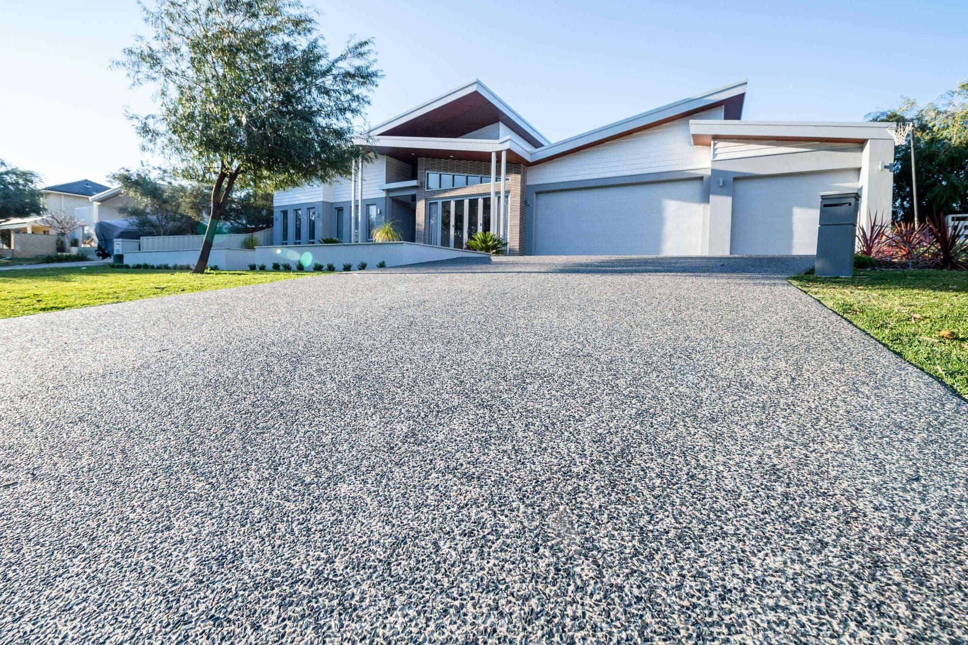 A modern house in Glendale, AZ boasts a large, sloped driveway crafted by expert concrete contractors, leading to a spacious three-car garage. The home features angular roof designs, extensive windows, and a well-maintained front lawn with trees and landscaping. The sky is clear, indicating a sunny day.