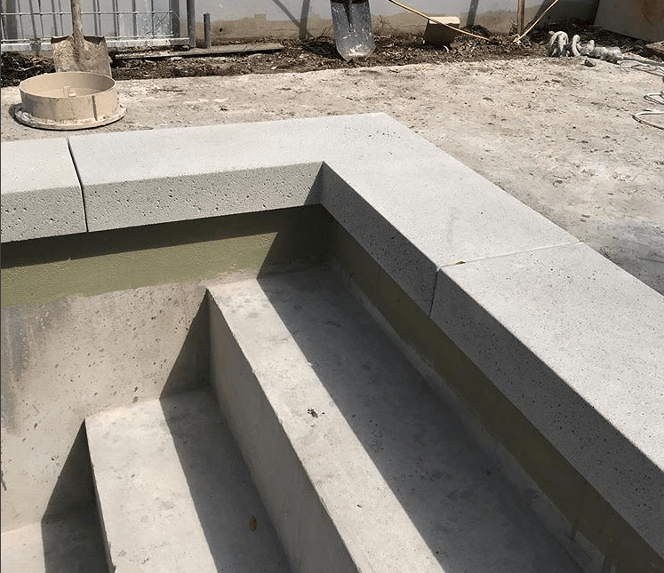 A close-up view of a concrete staircase under construction leading into what appears to be an unfinished pool. The surrounding area shows construction materials and tools, including a shovel and a plastic container. Some dirt and debris are visible on the ground, typical of work by West Valley Concrete, a trusted pool deck contractor in Glendale AZ.